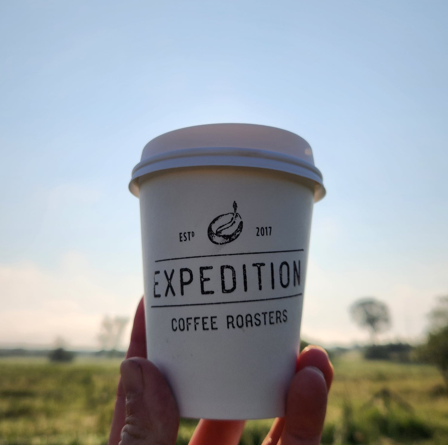 White paper cup with 'Expedition Coffee Roasters' branding held against a blurred natural background